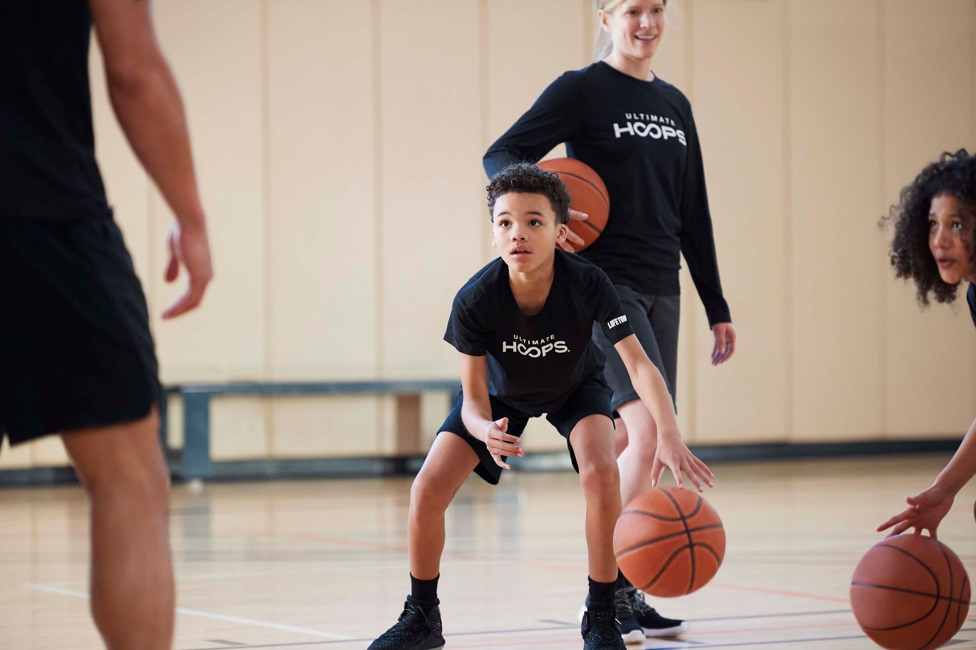 Children and adult playing basketball indoors, wearing black shirts with 'Ultimate Hoops' logo.