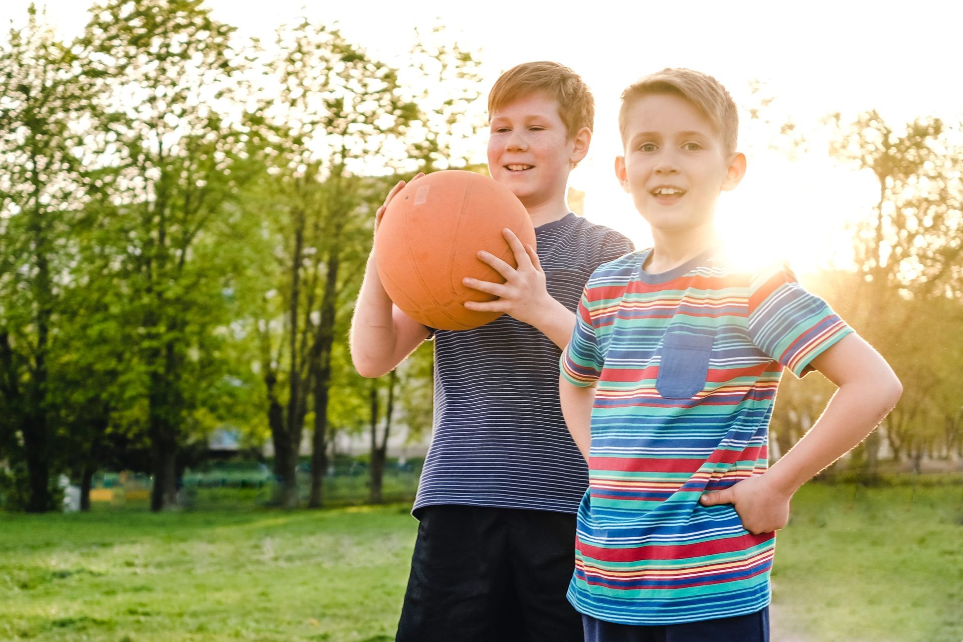 Two happy young boy friends or brothers standing on a rural sports field holding up a basketball with happy grins backlit by the glow of the setting sun